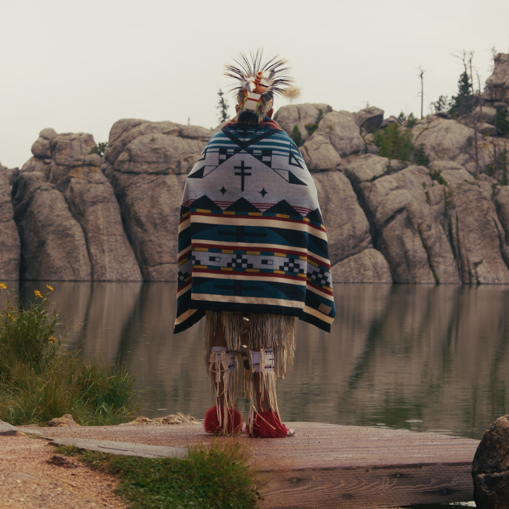Person wearing a patterned blanket and headdress standing on a wooden platform by a lake with rock formations in the background.